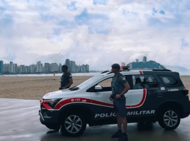 Imagem de uma viatura da Polícia Militar estacionada à beira-mar, com dois policiais fardados em pé ao lado. Ao fundo, há prédios altos, uma praia e um céu nublado.