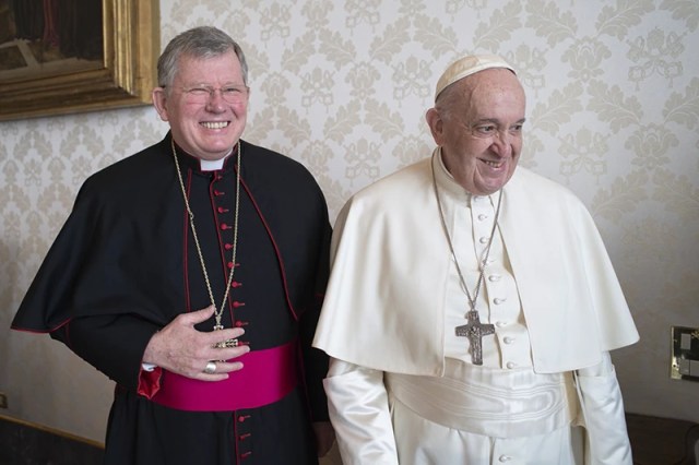 Papa Francisco sorri ao lado de um arcebispo vestido com batina preta e faixa roxa, ambos em um ambiente interno com parede decorada. A imagem pode estar relacionada à atuação de brasileiros no conclave e suas articulações na Igreja Católica.