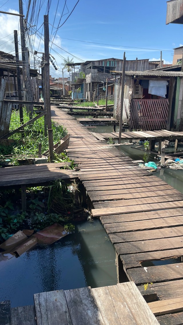 Comunidade ribeirinha em Belém com moradias precárias construídas sobre palafitas e passarelas de madeira acima de um igarapé poluído.