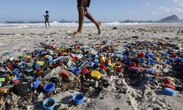 Tampas e resíduos plásticos espalhados pela areia de uma praia, com pessoas caminhando ao fundo, próximo ao mar.