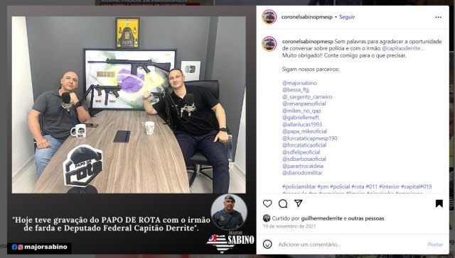 Coronel da PM SP participando de gravação do podcast “Papo de Rota” com outro policial, mesa com microfones e armas em exposição ao fundo.
